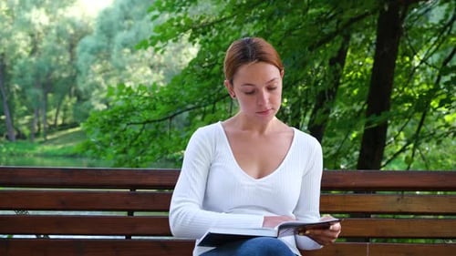 Young Woman Reading Book in Park By the River at Sunny Summer Day