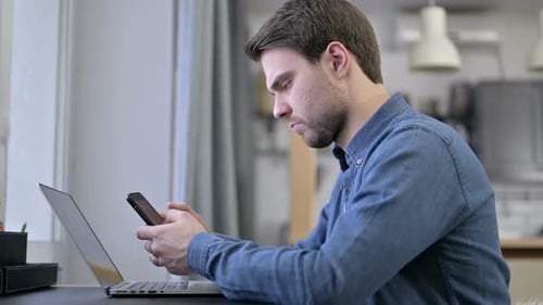 Man Using Smartphone and Laptop at Desk
