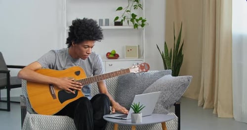 Young Adult Plays Guitar at Home Using Laptop