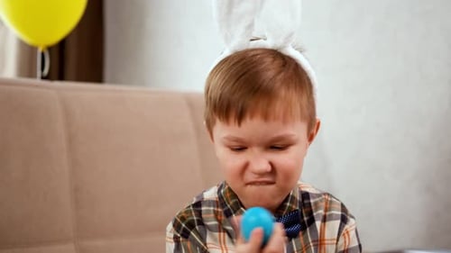 A Boy in Bunny Ears Holds a Blue Egg in His Hands and Depicts a Rabbit