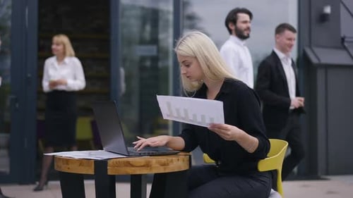 Concentrated Female Analyst with Paperwork Analyzing Emarket on Laptop As Colleagues Passing at