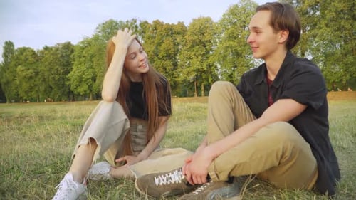 Positive Relaxed Teenage Couple Sitting on Summer Meadow Chatting and Laughing