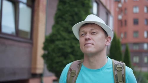 Young Man in Hat and Backpack Walks Around the City Camera Tracking