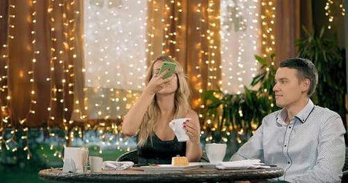 Man Looks at Girlfriend Taking Picture of Cappuccino in Cafe