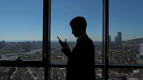 Silhouette of Young Businessman in Office Is Browsing Phone Near the Window with Panoramic City View