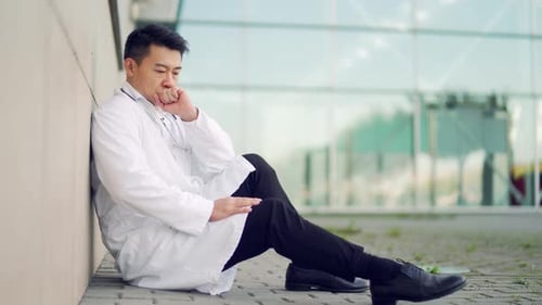 portrait stressful Asian doctor sitting or standing outside a hospital