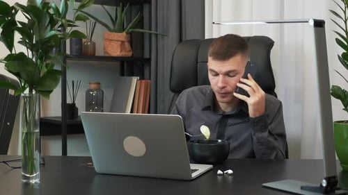 Businessman is eating in front of computer and speaking on smartphone at office desk