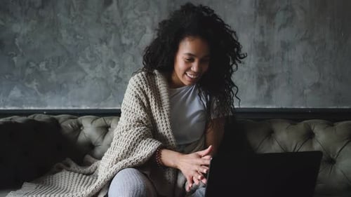 Junge attraktive afroamerikanische Frau, die Laptop für einen Videoanruf benutzt, der auf dem Sofa sitzt und