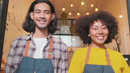 Two baristas at a cafe door, arms crossed, look at a camera with welcome smile.