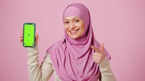 Happy Muslim Lady in Headscarf Holding Smartphone with Green Chroma Key Screen and Pointing at It