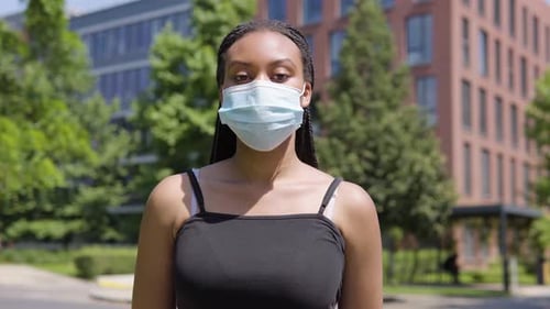 A Young Black Woman in a Face Mask Nods at the Camera - an Office Building and Trees