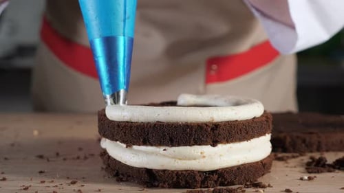 Pastry Chef Is Pouring Cream with Pastry Bag on Chocolate Sponge Cake, Making Cake in Bakery