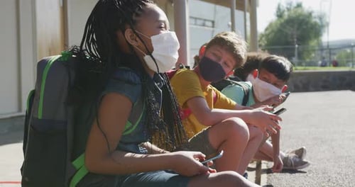 Grupo de niños que usan mascarillas y usan teléfonos inteligentes mientras están sentados juntos