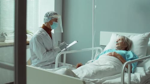 Doctor in Protective Uniform Talking with Senior Woman in Hospital