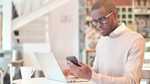 African Man Using Smartphone and Laptop in Cafe