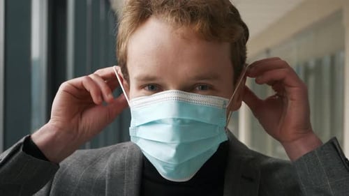 Close-up adult charming man wearing medical face mask, looking to camera on empty corridor backgroun