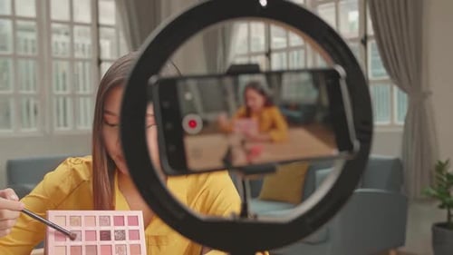 Mujer bloguera de belleza asiática con maquillaje en el estudio de su casa, grabando vídeo en un smartphone