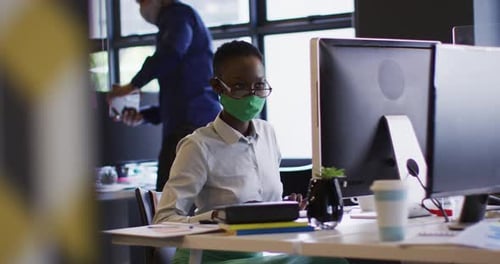 African american woman wearing face mask using computer while sitting on her desk at modern office