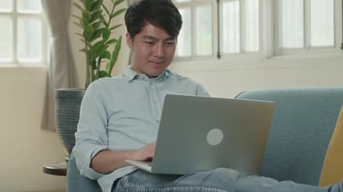 Hombre asiático usa una computadora portátil y sonríe en la sala de estar mientras está acostado en el sofá