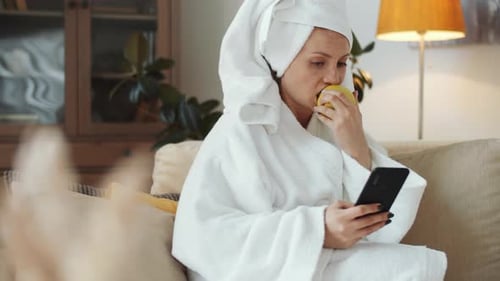 Woman in Robe Using Phone and Eating Apple