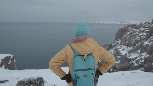 A Man in Bright Clothes and with a Backpack Stands on the Top of a Snowy Cliff and Admires the Sea