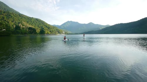 Two people in distance using stand up paddle board on lake amidst the mountains during early morning