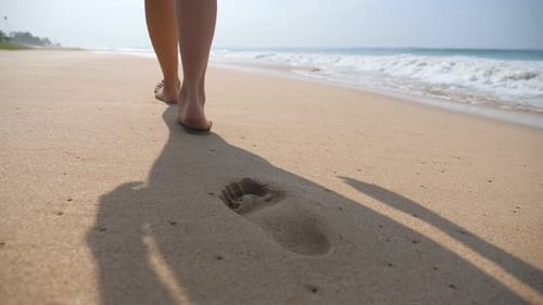 Female Feet Walking on Golden Sand at the Beach with Ocean Waves at Background. Legs of Young Woman