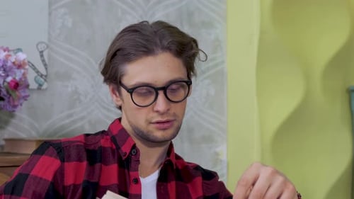 Close-Up Of A Young Man Sitting In A Bright Kitchen, Wearing Glasses And Taking A Book