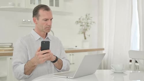Professioneller Mann mittleren Alters, der Smartphone und Laptop zu Hause benutzt