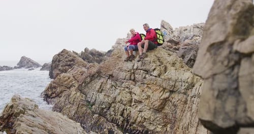 Senior hiker couple with backpacks sitting and talking on the rocks while hiking near sea shore.