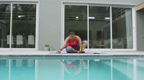 Happy african american plus size woman practicing yoga, using smartphone next to swimming pool