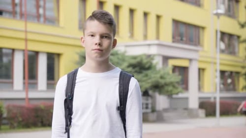 A Caucasian Teenage Boy Looks Seriously at the Camera a School in the Background