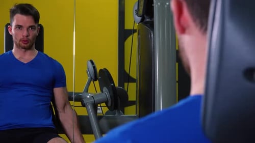 A Young Fit Man Trains on a Machine in a Gym - Closeup From Behind and View on the Mirror