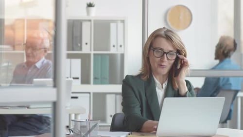 Smiling Middle-Aged Caucasian Lady Talking on Smartphone at Office Desk