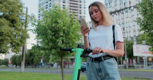 Pretty woman is using smartphone after ride on electric scooter in the urban city.