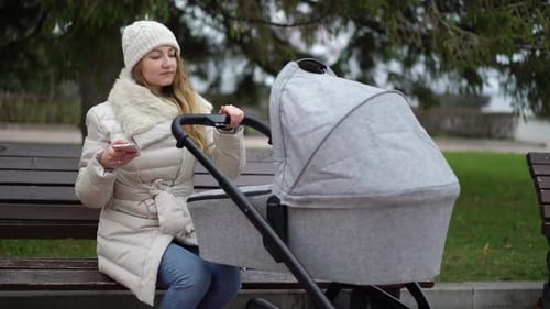 Happy Mother with Smartphone Using App. She Sitting on the Bench , Baby Sleeping in Pram