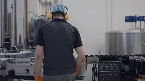 Back View Of An Operator Wearing Hairnet And Headphones Working In A Beer Factory