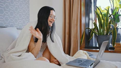 Junge süße Dame mit Brille winkt mit der Hand zum Laptop und begrüßt Freunde per Videoanruf