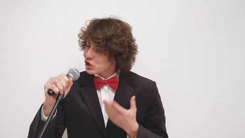 Un hombre de traje con pajarita roja sostiene un micrófono contra un fondo aislado