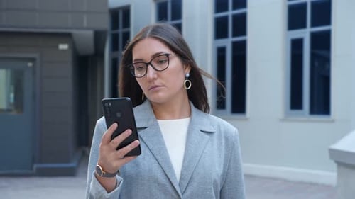Young Woman in Glasses and a Suit Walks Between Buildings Looking at the Phone Slow Motion