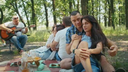 Two Young Couples Man and Woman Enjoy Their Vacation in the Forest While Playing the Guitar the