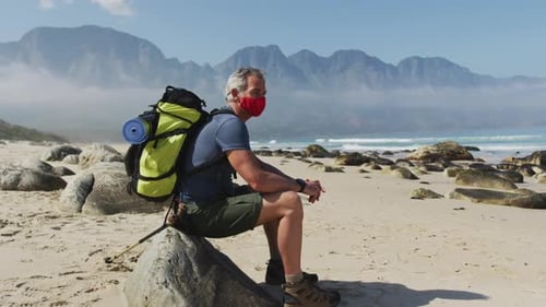 Senior hiker man wearing face mask with backpacks sitting on a rock while hiking on the beach.