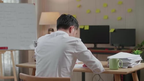 Back View Of Asian Man Writing Something On Paper While Working With Documents At The Office