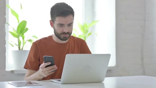 Man Using Smartphone and Laptop at Desk