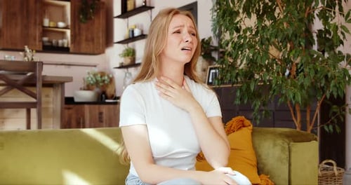 A young woman holds her hands over a sore throat. Painful neck and frowning