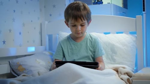 Portrait d'un petit garçon assis dans son lit la nuit et regardant une vidéo sur une tablette