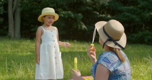 Funny Mom and Little Daughter Blow Soap Bubbles Sitting on the Lawn in the Park
