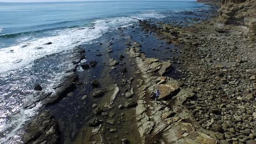 Aerial shot of young man running on a scenic rocky beach coastline.