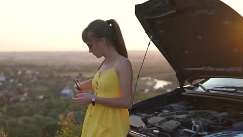 Young woman driver talking on mobile phone standing near her car with open hood