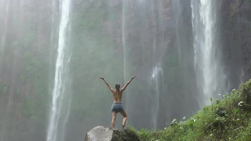 Woman Standing Under Tropical Waterfall with Arms Raised